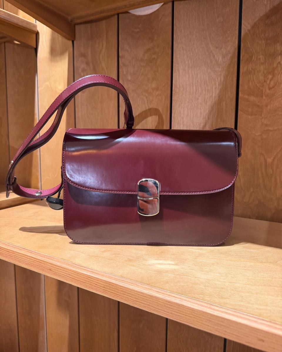 Bolsa de couro bordô com alça, apoiada em prateleira de madeira